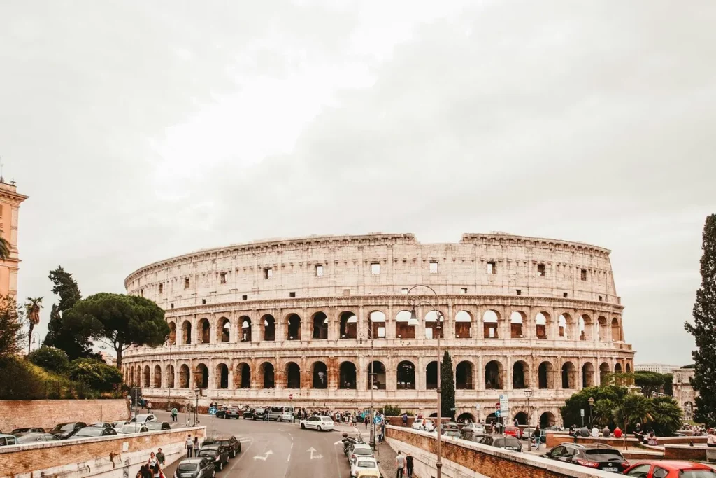 Vista frontal e panorâmica do Coliseu de Roma (Colosseum ou Anfiteatro Flaviano) em um dia nublado. O monumento de pedra clara é cercado por uma rua e carros, mostrando sua integração à paisagem urbana moderna.