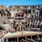 Vista aérea do interior do Coliseu de Roma com o piso da arena parcialmente reconstruído em madeira, revelando as ruínas do hipogeu (câmaras subterrâneas). Turistas se aglomeram ao redor e nos arcos superiores sob o céu azul.