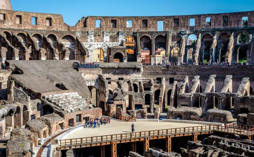 Vista aérea do interior do Coliseu de Roma com o piso da arena parcialmente reconstruído em madeira, revelando as ruínas do hipogeu (câmaras subterrâneas). Turistas se aglomeram ao redor e nos arcos superiores sob o céu azul.