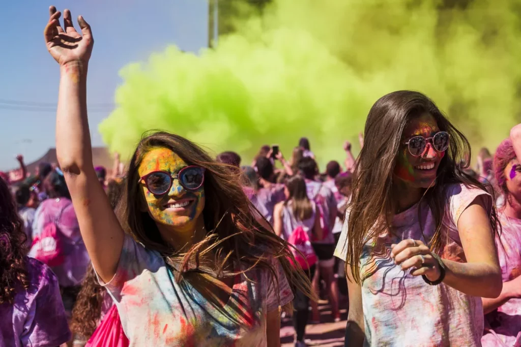 Duas mulheres com roupas e rostos coloridos sorriem e dançam durante o Holi Festival com fumaça verde ao fundo.