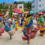 Apresentação de Maracatu Rural com brincantes em fantasias coloridas, chapéus ornamentados e cortejo popular em praça pública de Pernambuco.