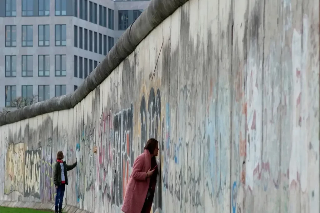Vista de uma longa seção remanescente do Muro de Berlim, altamente grafitada. Uma mulher em casaco rosa e um homem mais distante estão ao lado do muro, com prédios de escritórios modernos ao fundo.