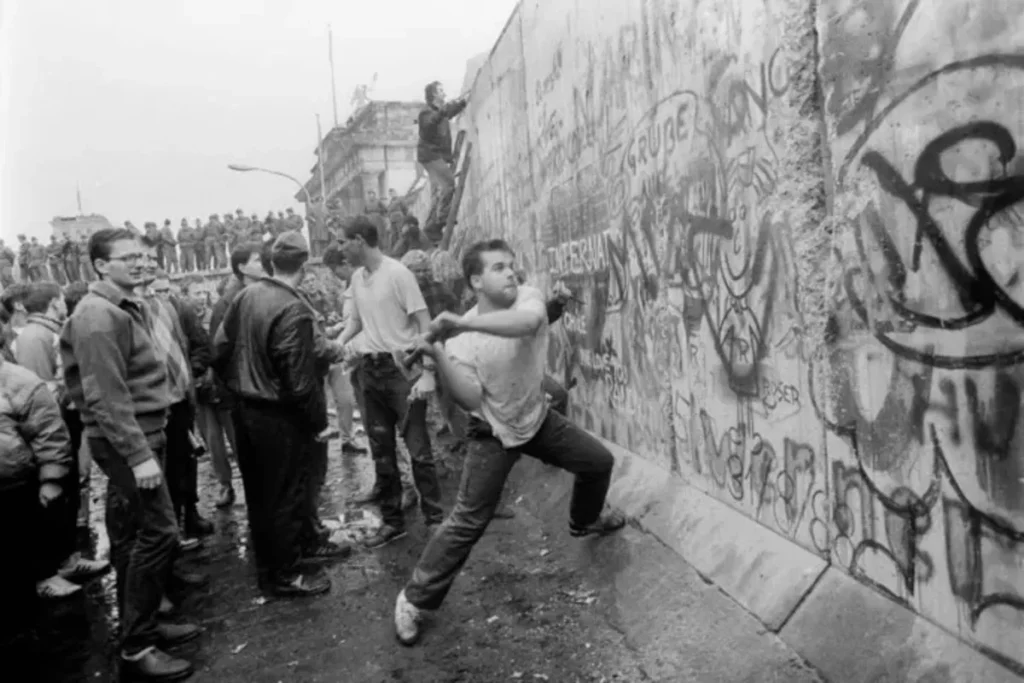 Fotografia histórica em preto e branco de um grupo de alemães ocidentais ou orientais celebrando a queda do Muro de Berlim em 1989. Um homem em primeiro plano martela o muro grafitado, enquanto outros observam e sobem no topo.