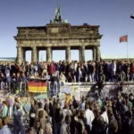 Fotografia histórica mostrando uma imensa multidão de pessoas em cima e ao redor do Muro de Berlim, celebrando em frente ao Portão de Brandemburgo, com bandeiras alemãs visíveis, no dia da queda em 1989.