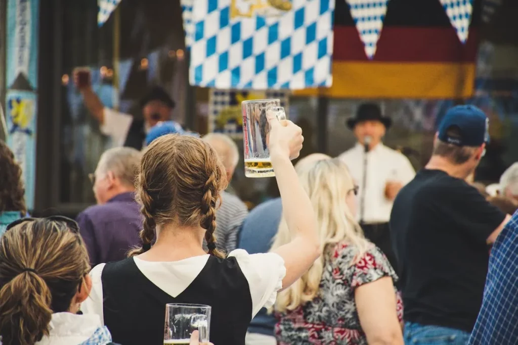 Mulher brinda com caneca de cerveja em festa Oktoberfest ao ar livre, cercada por pessoas e decoração típica alemã.