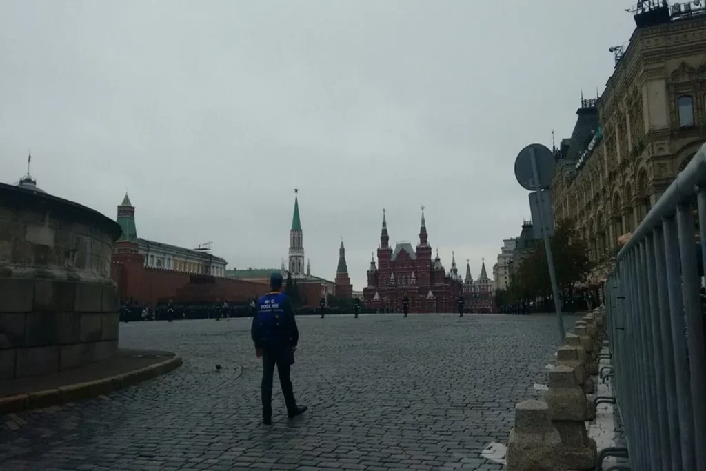 Vista de um dia nublado na Praça Vermelha em Moscou. Um segurança com colete azul (Policia Russa ou Segurança) está caminhando sobre o chão de paralelepípedos, com o Museu Histórico do Estado vermelho e o muro do Kremlin ao fundo.