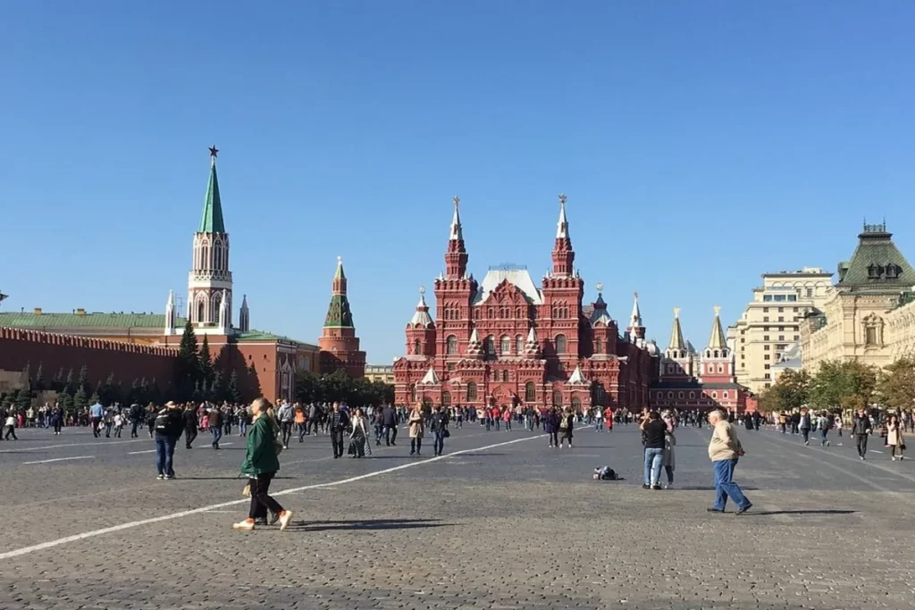 Vista diurna e ampla da Praça Vermelha em Moscou, mostrando o grande Museu Histórico do Estado (edifício vermelho central) e parte do muro do Kremlin com as torres e a bandeira russa ao longe, sob um céu azul-claro.