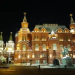 Explorando a Praça Vermelha em Moscou: do passado ao presente em uma viagem surreal Vista noturna da Praça Vermelha em Moscou, com o Museu Histórico do Estado (edifício vermelho) totalmente iluminado em tons de ouro e laranja, e o muro do Kremlin com torres iluminadas à esquerda, sob um céu preto.