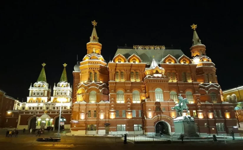 Vista noturna da Praça Vermelha em Moscou, com o Museu Histórico do Estado (edifício vermelho) totalmente iluminado em tons de ouro e laranja, e o muro do Kremlin com torres iluminadas à esquerda, sob um céu preto.