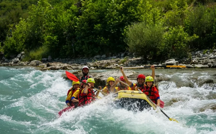 Grupo de pessoas praticando rafting em corredeiras, usando coletes salva-vidas e capacetes coloridos em bote inflável amarelo.