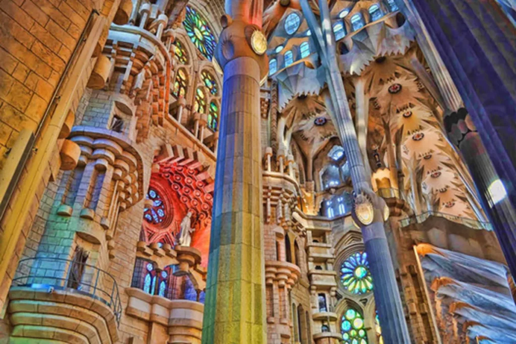 Vista interior de ângulo baixo e dramático da Basílica da Sagrada Família, mostrando a complexidade das colunas, as abóbadas e a luz colorida que entra pelos vitrais (azul, laranja, amarelo) de perto.