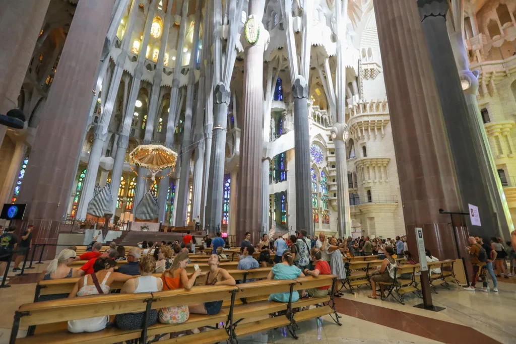 Vista interior da Basílica da Sagrada Família em Barcelona, mostrando as colunas gigantes de pedra, vitrais coloridos e o teto alto, com turistas sentados nos bancos de madeira.