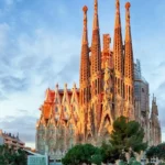 Vista exterior da Basílica da Sagrada Família em Barcelona, mostrando as torres altas da Fachada da Natividade com andaimes e pedra alaranjada, banhada pela luz quente do pôr do sol sob um céu azul e nublado.