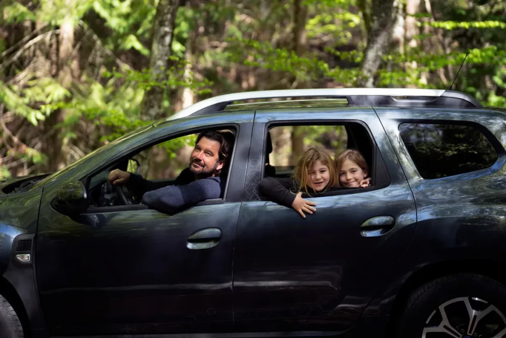 Pai e duas filhas em um carro, sorrindo e aproveitando a paisagem durante uma viagem.