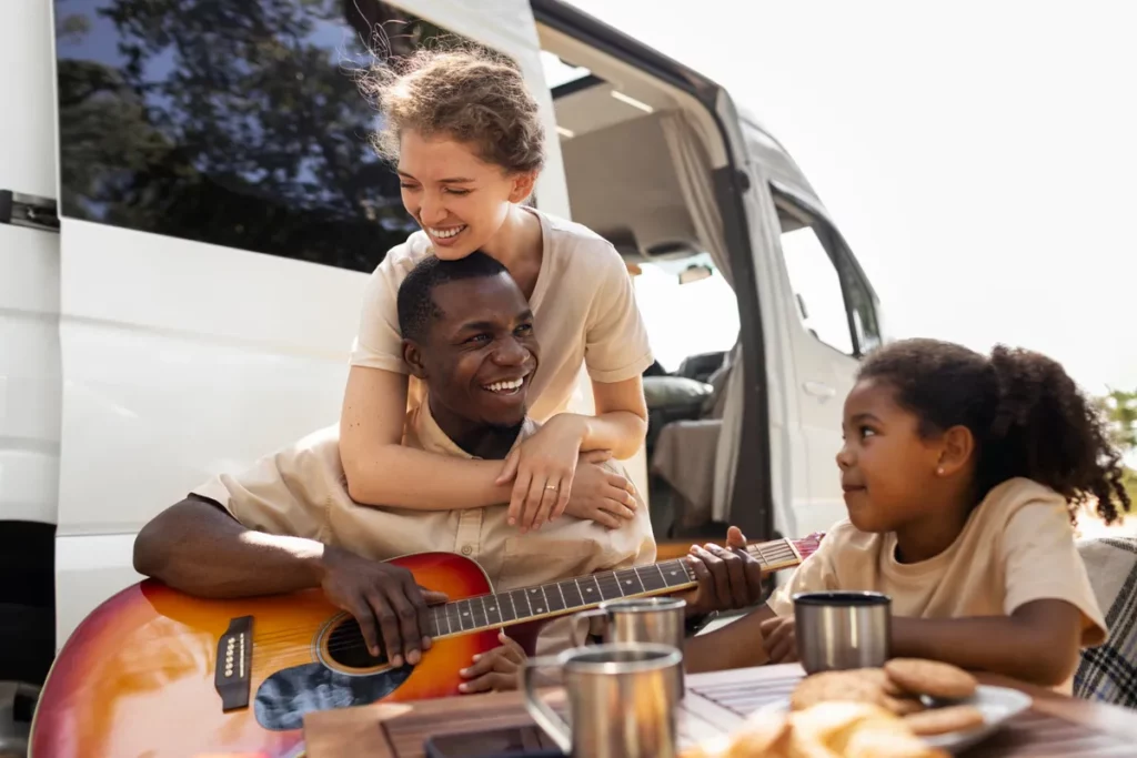 Família sorridente em frente a uma van alugada, pai tocando violão, mãe abraçando-o e filha curtindo o momento em uma mesa ao ar livre.