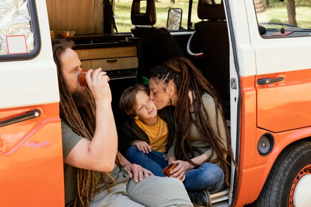 Casal com filho pequeno sentado na porta de uma van de família laranja e branca, curtindo juntos um momento de carinho em viagem.