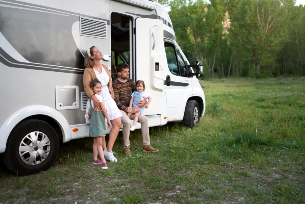 Família com duas crianças em frente a uma van motorhome branca alugada, parada em meio à natureza durante viagem.