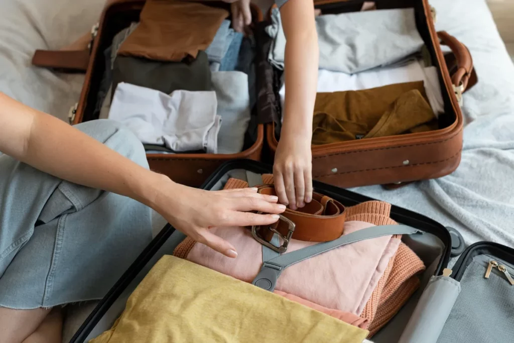 Mãos de um adulto e uma criança arrumando duas malas de viagem abertas, colocando roupas e um cinto. Cena de organização de bagagem de mão para viajar.