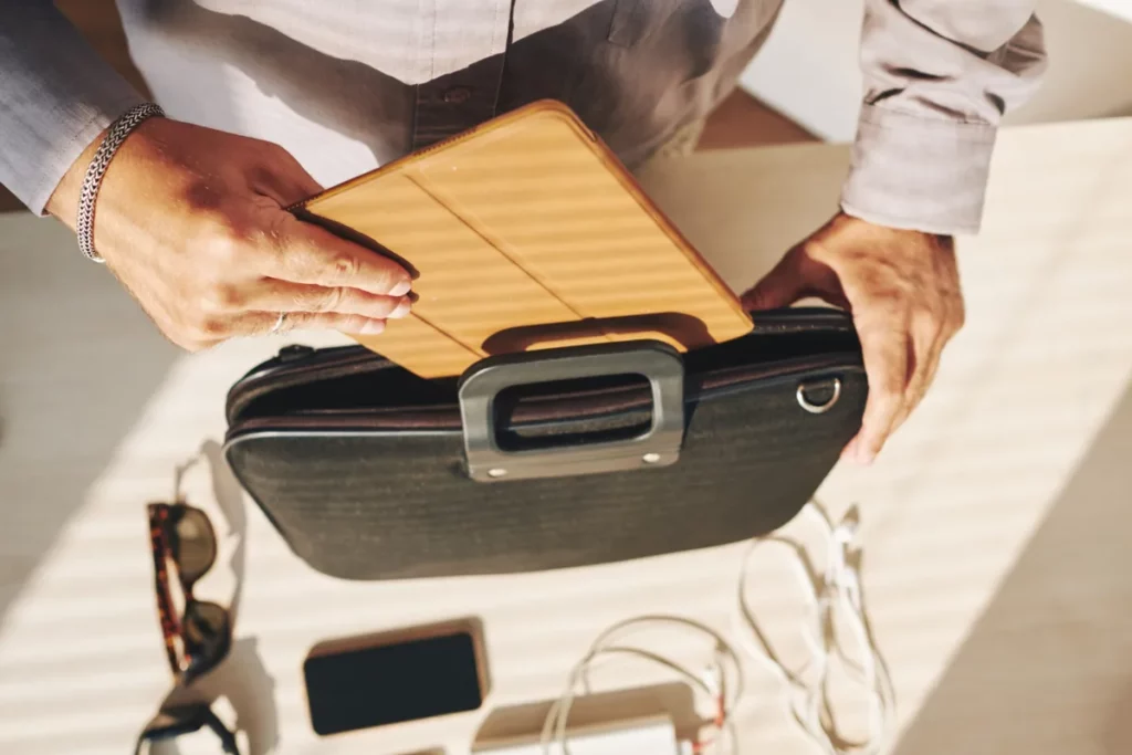 Mãos de uma pessoa arrumando uma bolsa de bagagem de mão, colocando um tablet em uma capa marrom. Acessórios de viagem, como óculos de sol e celular, estão sobre a mesa.