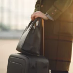 Pessoa com casaco xadrez segurando uma mala de rodinhas preta e uma bolsa de couro preta. Cena de viagem com bagagem de mão em um aeroporto.