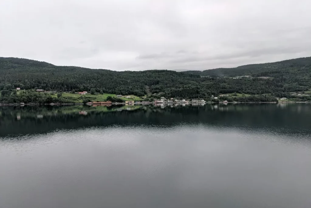 Vista panorâmica de um fiorde escandinavo em um dia nublado, com montanhas verdejantes cobertas por florestas, uma pequena aldeia de casas coloridas na margem e a superfície da água escura e calma refletindo a paisagem.