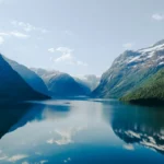 Vista panorâmica de um fiorde profundo e azul-turquesa, cercado por montanhas íngremes e cobertas por florestas verdes e picos com neve. O reflexo das montanhas na água calma é simétrico sob um céu azul-claro com nuvens brancas.