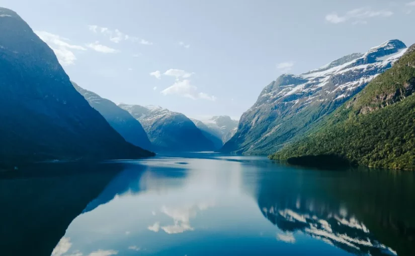 Vista panorâmica de um fiorde profundo e azul-turquesa, cercado por montanhas íngremes e cobertas por florestas verdes e picos com neve. O reflexo das montanhas na água calma é simétrico sob um céu azul-claro com nuvens brancas.