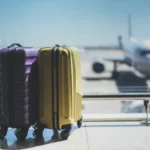 Duas malas de viagem, uma roxa e uma amarela, no saguão de aeroporto com avião ao fundo.