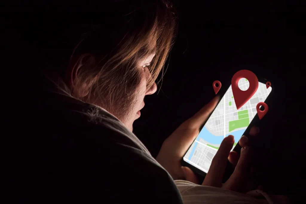 Perfil de uma mulher em um ambiente escuro, iluminada apenas pela tela de um smartphone que exibe um mapa com marcadores de localização (pinos vermelhos) flutuantes, ilustrando a dependência de GPS ou mapas offline à noite.