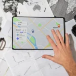 Mão tocando um tablet que exibe um mapa digital e informações de restaurante, com mapas de papel espalhados, óculos e uma câmera DSLR ao redor, ilustrando o planejamento de rota e o uso de mapas offline.