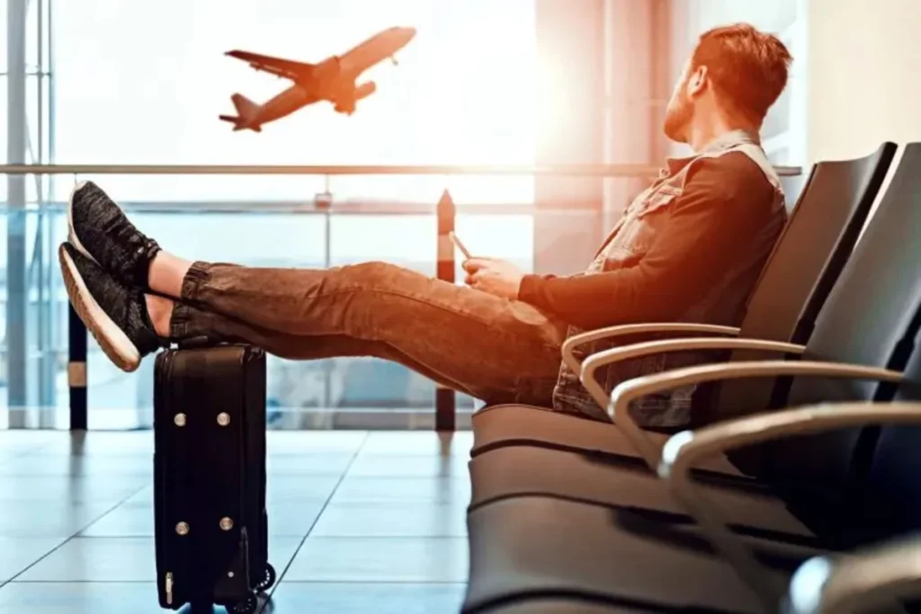 Homem casualmente vestido sentado em assentos de aeroporto com os pés apoiados em uma mala, olhando para um avião decolando pela janela, sugerindo uma espera tranquila ou relaxamento pré-viagem.