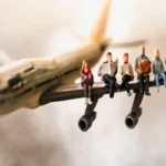 Miniaturas de seis pessoas sentadas na asa de um avião de brinquedo em pleno voo, uma metáfora visual para o overbooking e o excesso de passageiros, com foco na figura central lendo um jornal.