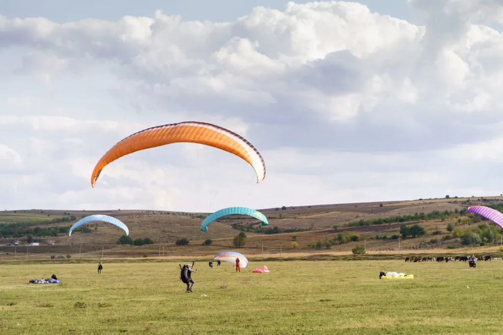 Foto de um campo com várias pessoas e parapentes. Três parapentes coloridos estão voando no céu, enquanto outros estão no chão sendo preparados para decolar.