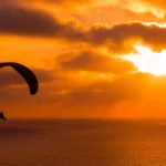 Silhueta de um parapente voando contra um pôr do sol alaranjado e avermelhado sobre o mar.