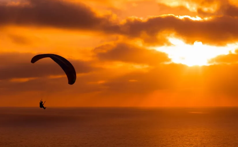 Silhueta de um parapente voando contra um pôr do sol alaranjado e avermelhado sobre o mar.