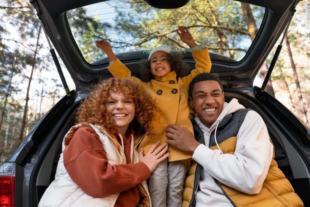 Família diversa e feliz sentada no porta-malas de um carro, aproveitando uma pausa em uma road trip.