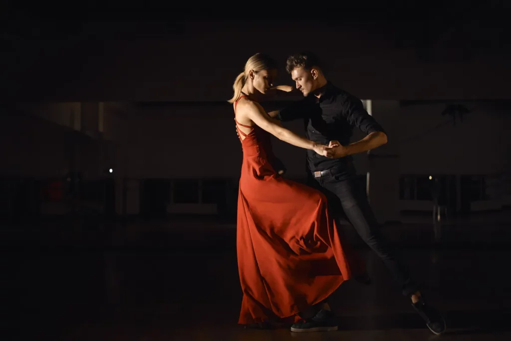 Casal dançando tango argentino em ambiente escuro, com a mulher de vestido vermelho em movimento.