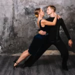 Casal dançando tango argentino em pose elegante, com fundo de parede cinza e atmosfera artística.