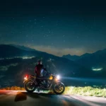 Motociclista parado com sua moto observando o céu estrelado e as luzes da cidade nas montanhas.