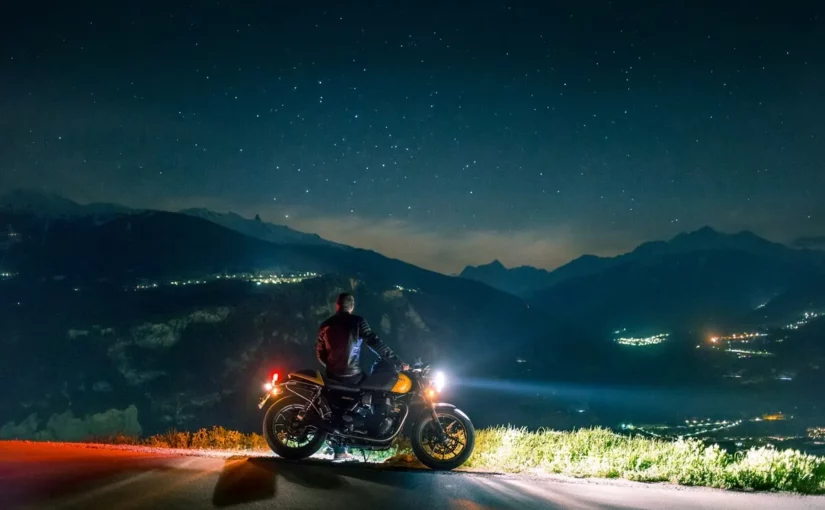 Motociclista parado com sua moto observando o céu estrelado e as luzes da cidade nas montanhas.