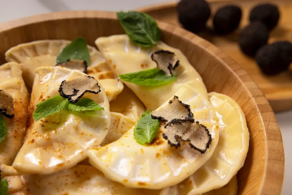 Close-up de um prato de Pierogi (bolinhos de massa recheados, de origem polonesa) servido em uma tigela de madeira. Os Pierogi estão cobertos com um molho cremoso, salpicados com pimenta e finas raspas de trufa preta, e decorados com folhas de hortelã fresca, destacando uma versão gourmet.
