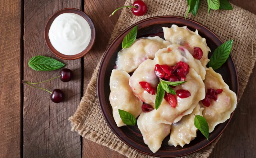 Vista de cima de um prato de Pierogi (bolinhos de massa recheada) na versão doce, com o recheio de cerejas (ou frutas vermelhas) aparente, servido em um prato de barro marrom. O prato está decorado com folhas de hortelã e acompanhado por uma tigela de creme azedo (sour cream) e cerejas frescas em um fundo rústico de madeira.