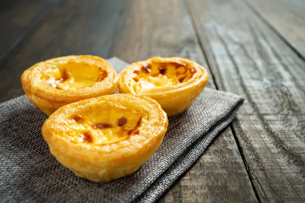 Três Pastéis de Nata com recheio de creme de ovo, casca folhada e marcas de caramelo, sobre um guardanapo cinza em mesa de madeira escura, culinária portuguesa.