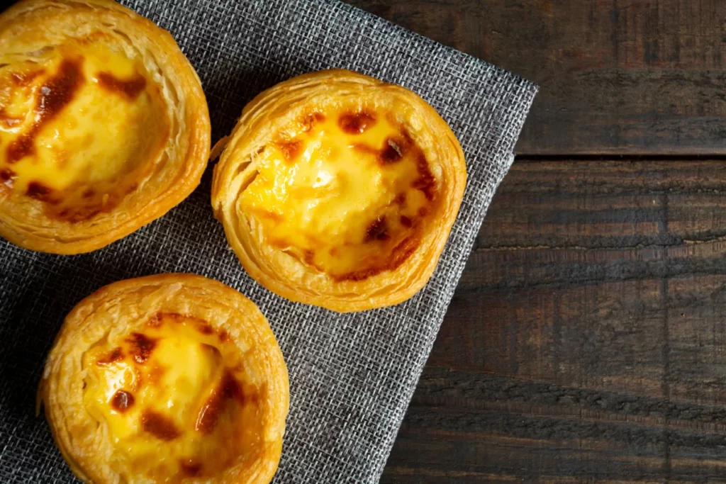 Três Pastéis de Nata com recheio cremoso e casca folhada crocante, sobre tecido cinza em fundo de madeira escura, culinária portuguesa.