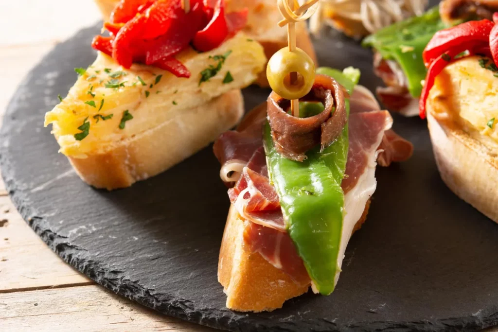 Tapas espanholas (Montaditos) de presunto cru, pimenta verde, aliche e azeitona em fatia de pão, sobre prato de ardósia escura.