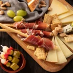 Tábua de frios e queijos com presunto cru em grissini, queijo azul, azeitonas, picles e amêndoas, ideal para servir como tapas espanholas.
