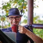 Homem em uma rede, tomando café e com seu laptop