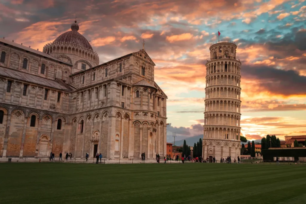 Torre de Pisa inclinada ao lado da catedral na Praça dos Milagres durante o pôr do sol, com visitantes caminhando pelo gramado