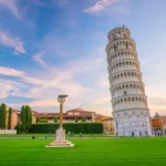 Torre de Pisa inclinada vista de frente na Praça dos Milagres em dia claro, com gramado e construções históricas ao redor