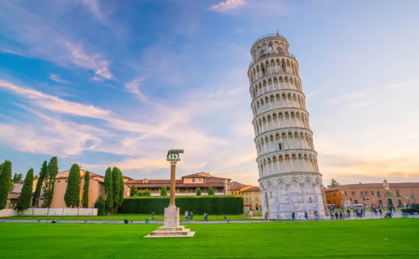 Torre de Pisa inclinada vista de frente na Praça dos Milagres em dia claro, com gramado e construções históricas ao redor